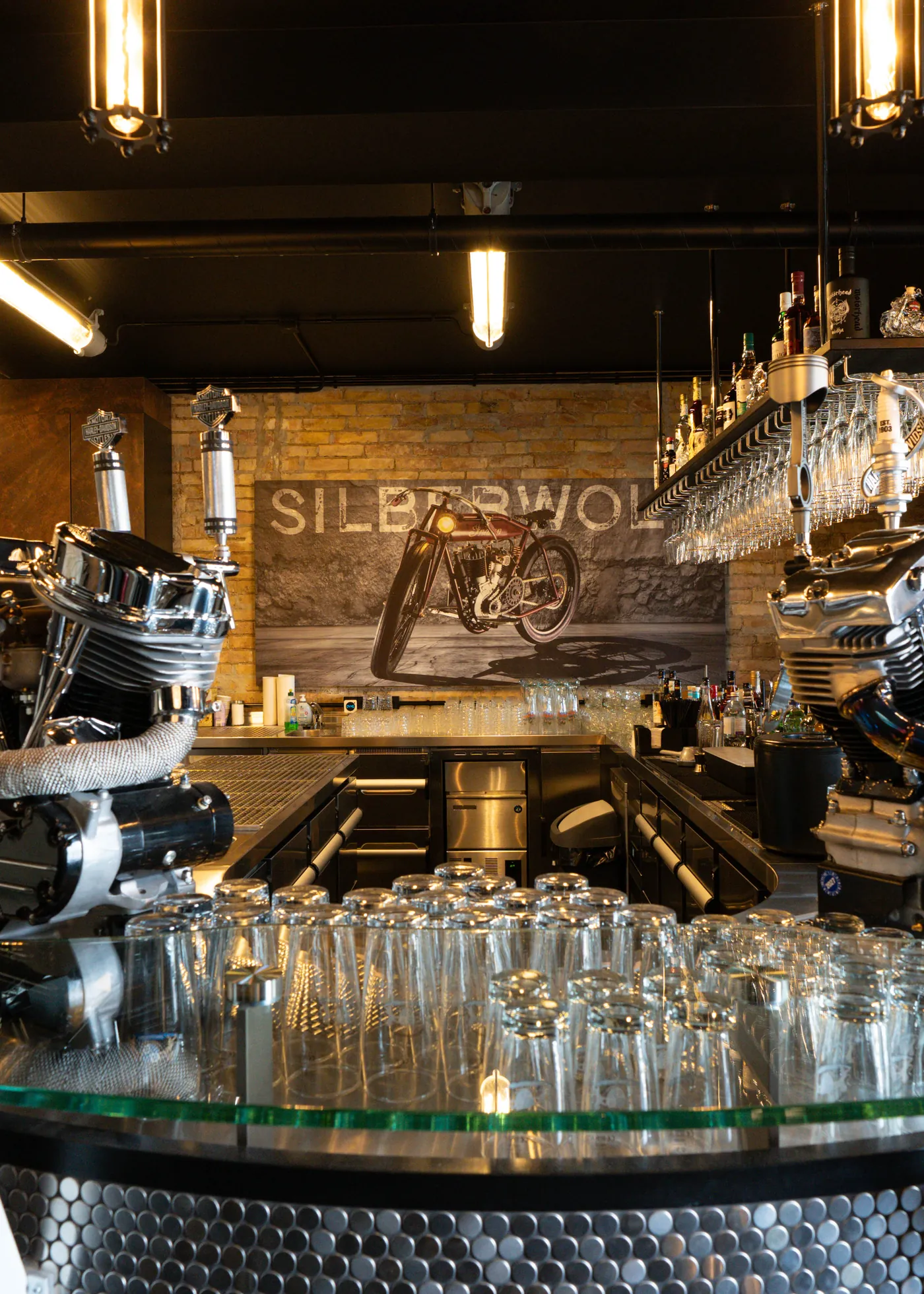 Two chromed engine blocks on the glass top of the counter, rows of glasses underneath