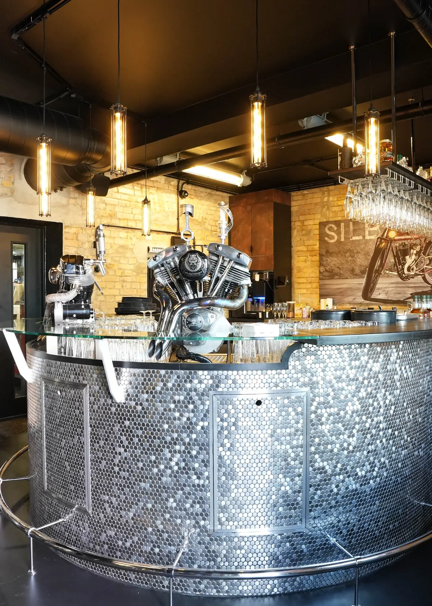 The bar of the Silberwolf Biker Bar: a curved counter of studded stainless steel with a chromed V-twin engine on top, brick wall with the Silberwolf lettering behind it