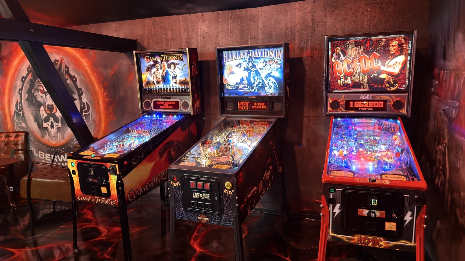 Three lit pinball machines side by side on the barn's lava floor, the wolf crest on the wall behind